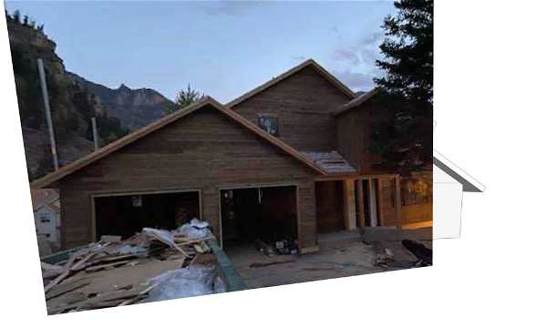 A partially constructed house with wooden exterior walls and a driveway cluttered with construction materials. The house features a two-car garage and is set against a backdrop of trees and possibly a mountain. The sky appears to be at dusk or dawn.