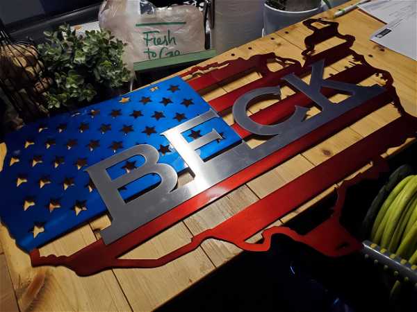 A metal cutout in the shape of the United States is placed on a wooden table. It is painted with a blue field of stars and red and white stripes, resembling the American flag. The word "BECK" is prominently displayed across the cut