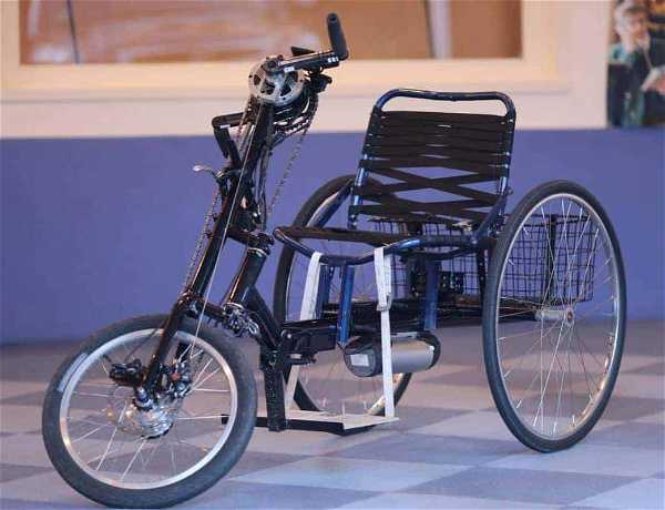 A three-wheeled handbike with a black frame, large rear wheels, and a smaller front wheel. The bike has a seat with back support and handlebars for steering, designed for individuals with mobility impairments. The handbike is positioned on a