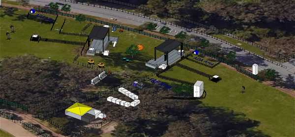 Aerial view of a festival layout featuring various structures and paths. The green field area includes several tents, stages, walkways, and fenced sections. Portable toilets are scattered throughout the premises, and there are trees and a road visible in the background.