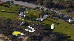Aerial view of a festival layout featuring various structures and paths. The green field area includes several tents, stages, walkways, and fenced sections. Portable toilets are scattered throughout the premises, and there are trees and a road visible in the background.